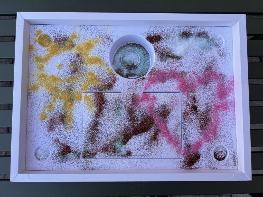photo of a macbook pro box with an apple airtag inside it. The inside of the box is covered in glitter of various colors. There is a yellow sun and a pink heart drawn with glitter
