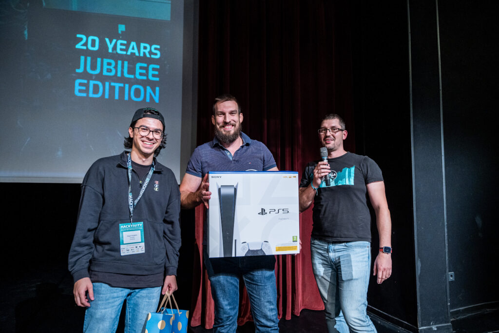 Photo of the Hacktivity speedhacking CTF final with both finalists and the organizer. The winner holding a PS5 box, and the organizer is holding the microphone.